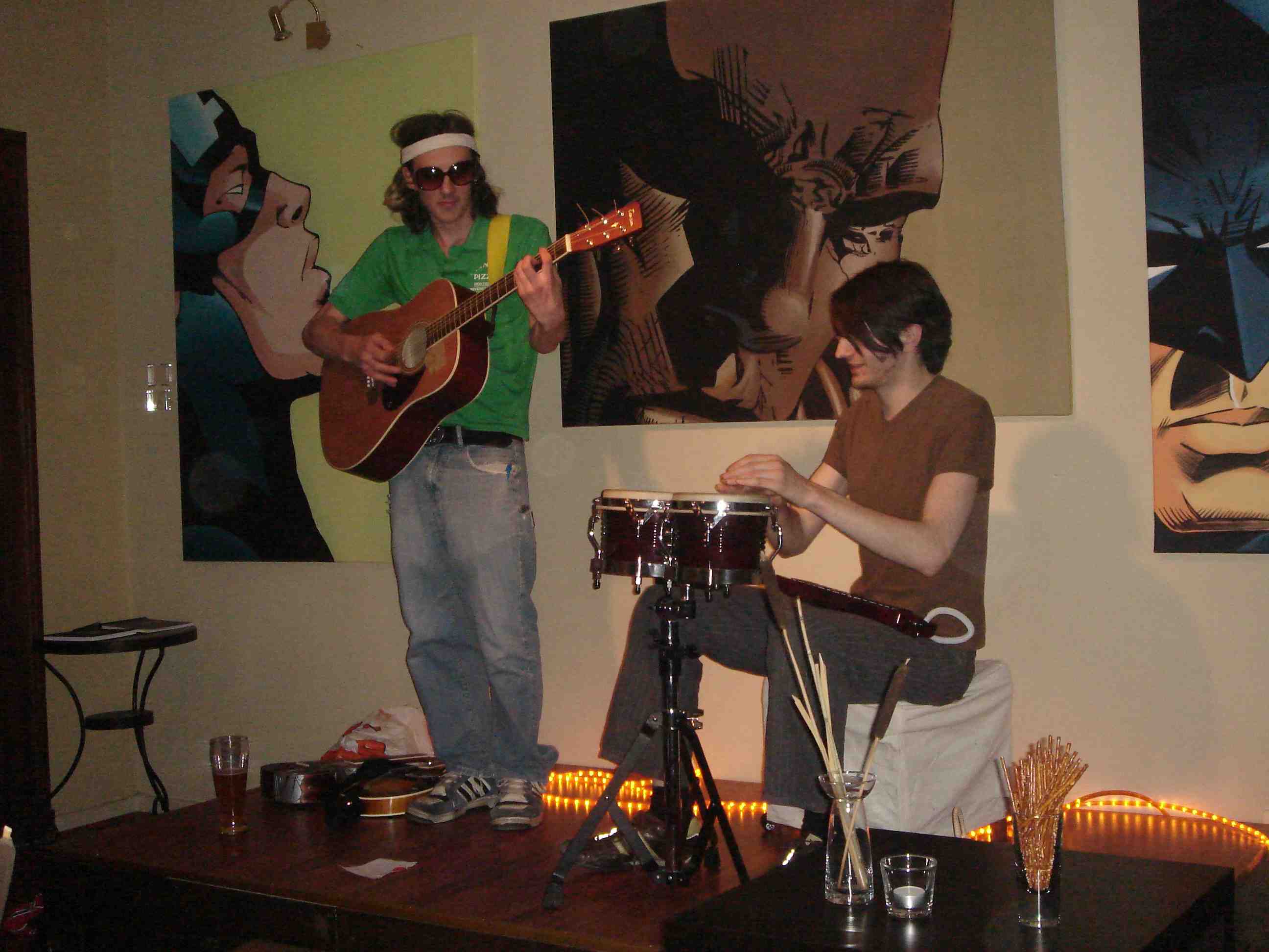 Two guys playing music on a tiny stage in front of somewhat homoerotic paintings of superheros